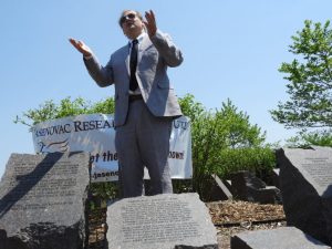 A person in a suit stands outdoors among engraved stones, gesturing expressively.