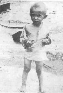 A young child stands barefoot on dirt, wearing a tattered shirt and shorts.