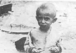 A young child stands barefoot on dirt, wearing a tattered shirt and shorts.